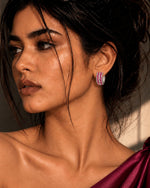 A close-up portrait of a woman wearing a small, vibrant pink and purple striped hoop earring, styled with a dark satin one-shoulder dress under dramatic warm lighting.