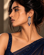 A woman in profile wearing a deep navy blue embellished sari and matching dangling earrings with blue and white stones, set against a softly lit neutral background.