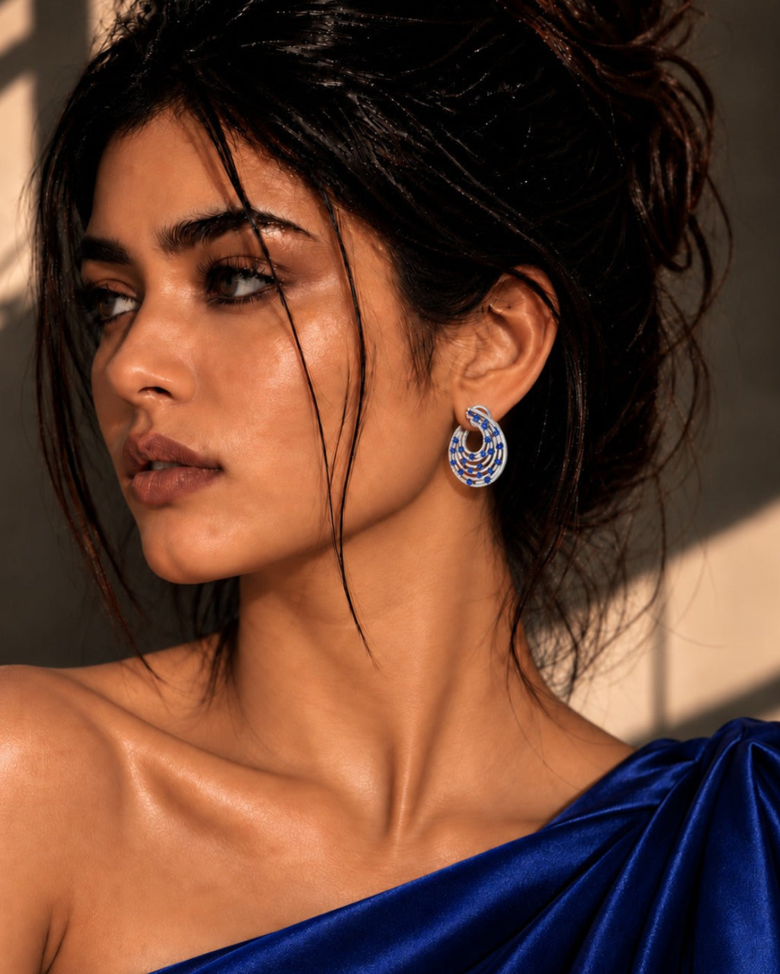 A close-up portrait of a woman wearing a blue one-shoulder dress and a blue-and-white patterned spiral earring, set against a warm-toned background with dramatic shadows.