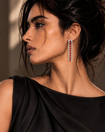 A woman in a black draped dress shown in profile, wearing a long drop earring with red stones and silver-toned settings. The background is a softly lit warm brown studio setting.