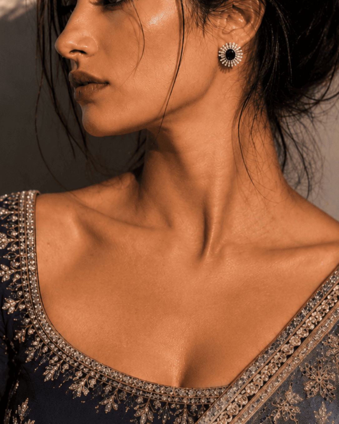 A woman in profile wearing a dark navy blue embroidered blouse and matching sheer draped fabric with ornate gold floral detailing, photographed in warm directional light against a textured wall.