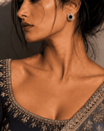 A woman in profile wearing a dark navy blue embroidered blouse and matching sheer draped fabric with ornate gold floral detailing, photographed in warm directional light against a textured wall.
