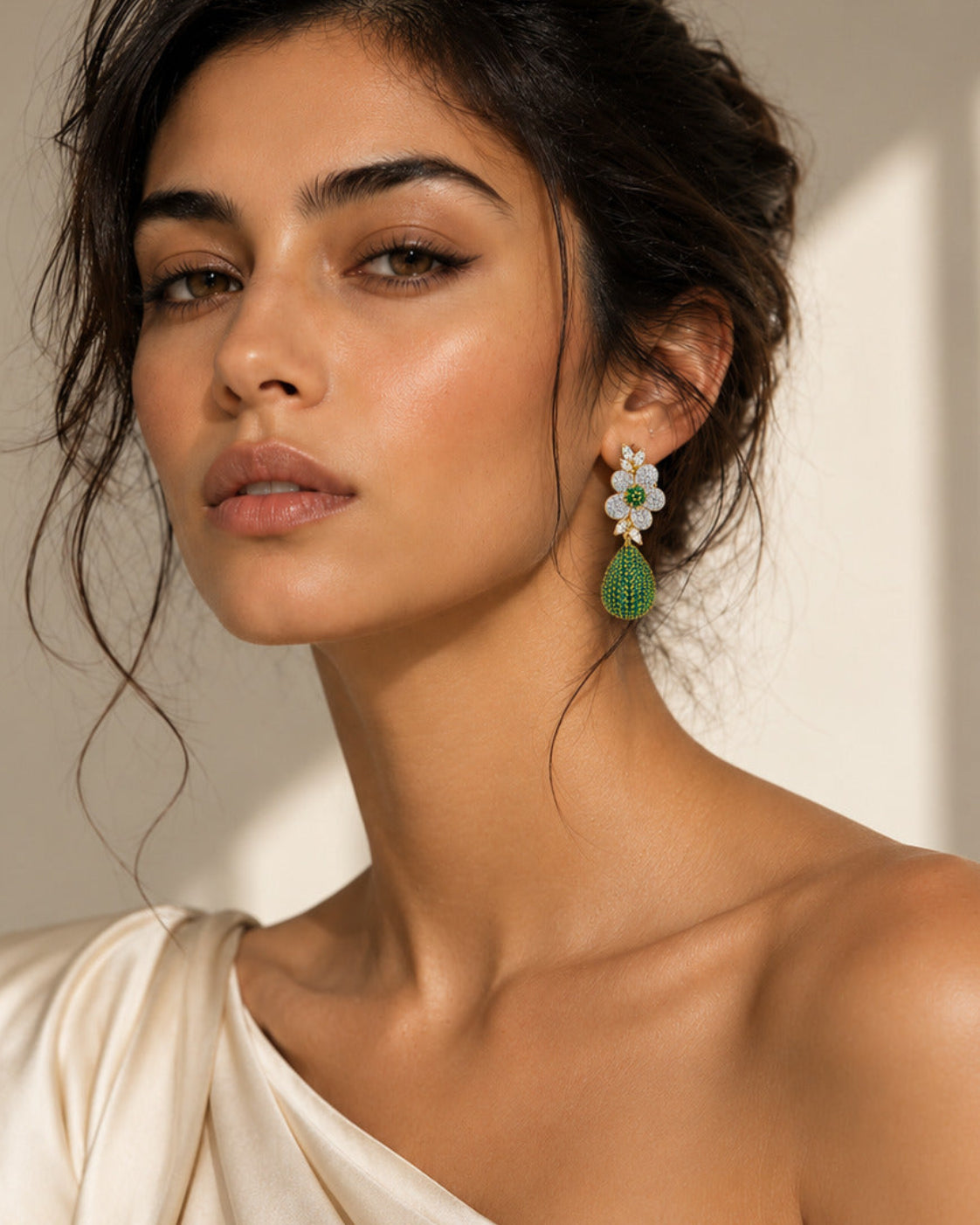 A close-up portrait of a woman wearing a dangling earring with a white floral cluster and a green teardrop-shaped stone, paired with a draped off-white top against a softly lit beige background.