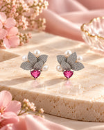 A pair of silver-toned butterfly earrings with pavé-set stones, white pearl accents, and a pink heart-shaped crystal at the center, displayed on a beige stone slab with pink flowers.