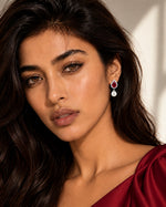 A close-up portrait of a woman wearing a teardrop-shaped drop earring with a red top stone and a white teardrop pendant, paired with a dark red satin top.