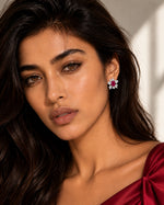 A close-up portrait of a woman with long dark hair wearing a small flower-shaped earring with a pink gemstone and white stones, against a softly lit neutral background.