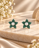 A pair of flower-shaped stud earrings with sparkling green pavé settings and white pearl centers, displayed on a beige marble slab with soft-focus floral props.
