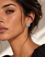 A close-up of a woman wearing a silver-toned, flower-shaped stud earring with clear stones. The earring is shown on her earlobe against a soft, warm-toned neutral background.