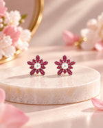 A pair of flower-shaped stud earrings with deep pink gemstone petals and white pearl centers, displayed on a round marble pedestal with soft pink floral props in the background.
