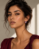 A close-up portrait of a woman wearing a small purple flower-shaped stud earring with a silver-toned center, paired with a deep burgundy draped top against a softly lit beige background.