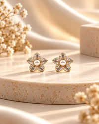 A pair of gold flower-shaped stud earrings with central white pearls and pavé-set clear stones, displayed on a beige stone pedestal with soft cream fabric and dried floral decor.