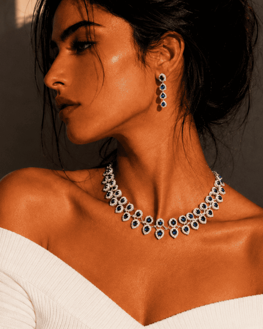 A woman in a white off-the-shoulder top models a sparkling silver-toned necklace and matching drop earrings with dark blue stones, set against a warm, shadowed beige wall.