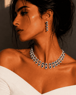A woman in a white off-the-shoulder top models a sparkling silver-toned necklace and matching drop earrings with dark blue stones, set against a warm, shadowed beige wall.