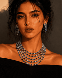 A woman wearing a multi-strand silver beaded necklace and matching drop earrings, photographed in dramatic warm lighting against a beige wall with strong shadows.