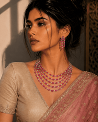 A woman wearing a pink saree with gold embroidery and a beige blouse, styled with a multi-strand pink gemstone necklace and matching dangling earrings.