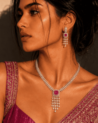 A woman wearing a silver-toned necklace and matching drop earrings with pink gemstones, styled with a magenta embroidered traditional garment against a warm beige background with dramatic shadows.