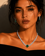 A woman with dark hair styled in a messy bun wears a sparkling diamond necklace with a central emerald-cut green gemstone and matching drop earrings, paired with a black off-the-shoulder top. The photo is taken in warm, dramatic golden-hour lighting with strong shadows against a beige wall.