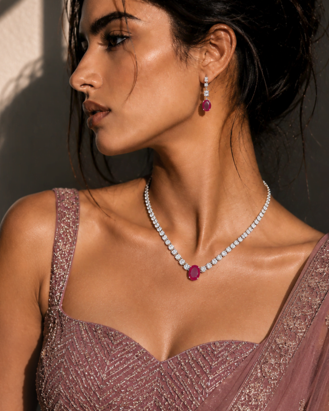 A woman in profile wearing a sparkling mauve embellished blouse and draped sari, styled with a matching pearl necklace featuring a deep pink gemstone pendant and matching dangling earrings.