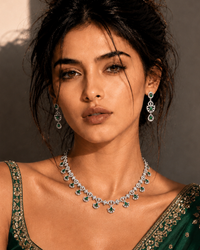 A woman wearing a dark green embroidered sari and matching jewelry, including a necklace and earrings, posed against a warm-toned wall with dramatic shadows.