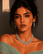 A woman with dark hair styled in a bun wears a sparkling silver-toned diamond necklace and matching drop earrings, paired with a sage green off-the-shoulder top. She is posed against a warm beige wall with dramatic shadows.