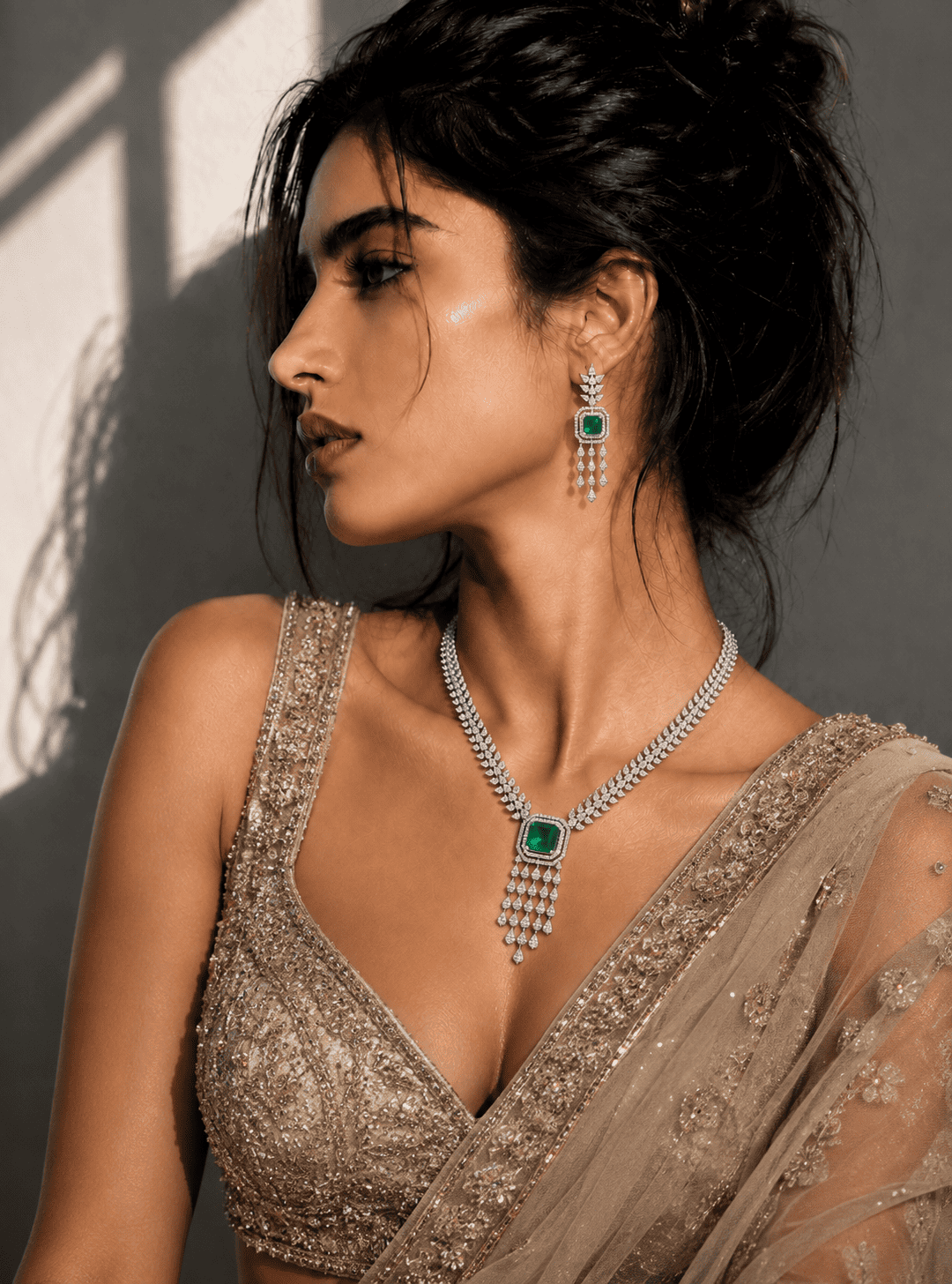 A woman in profile wearing an ornate silver-toned necklace with a green stone pendant and matching drop earrings, paired with a heavily embellished beige bridal outfit against a gray wall with dramatic shadows.