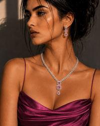 A woman in a shiny magenta satin dress models a diamond necklace with a pink gemstone pendant and matching drop earrings against a shadowed wall.