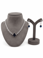 


A jewelry set displayed on gray velvet stands, featuring a silver-tone necklace with a dark blue teardrop centerpiece and matching drop earrings with blue oval stones and silver pavé details, set against a plain white background.
