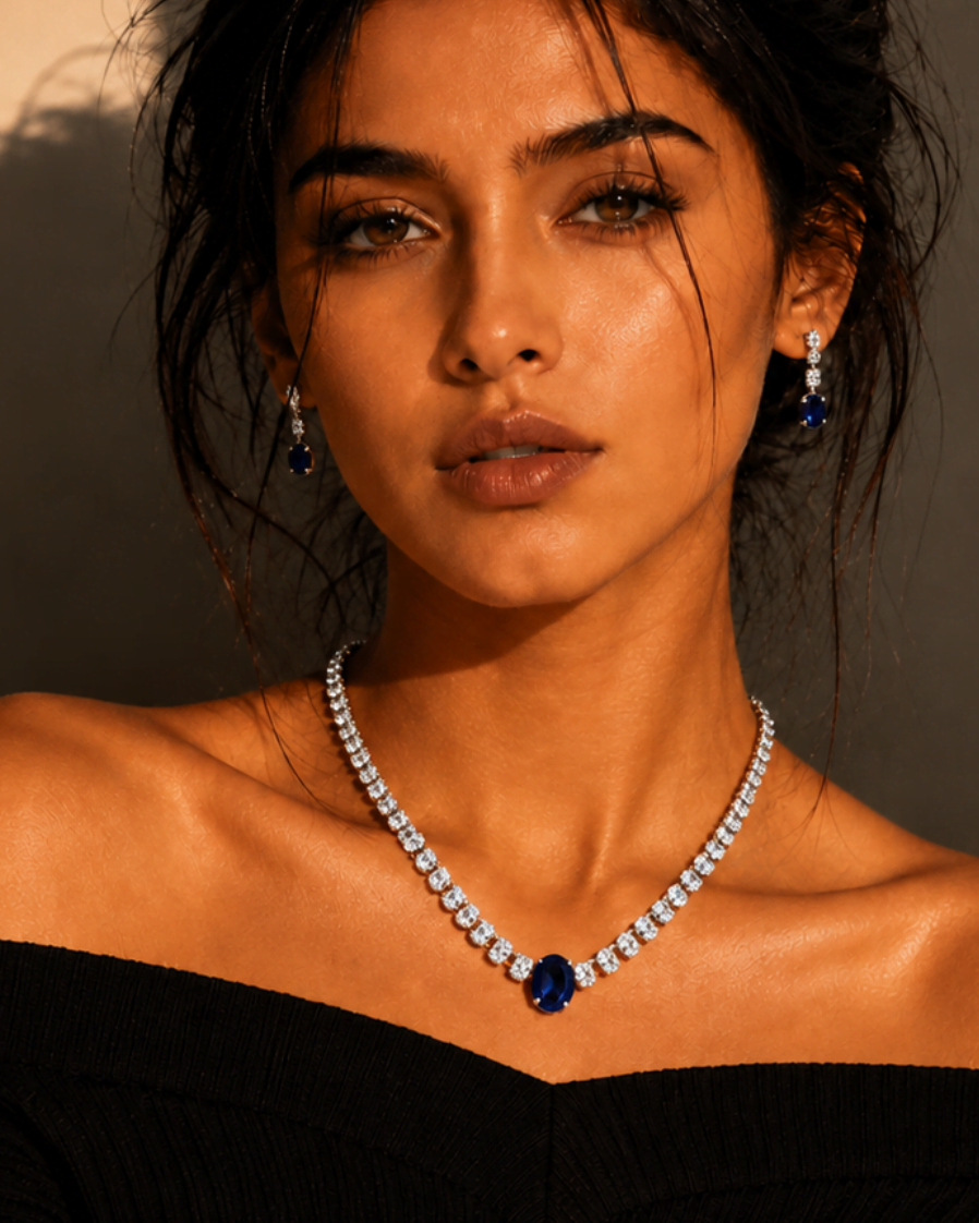 A woman wearing a sparkling diamond necklace with a dark blue oval center stone and matching drop earrings, styled with a black off-the-shoulder top against a wall with dramatic warm shadows.